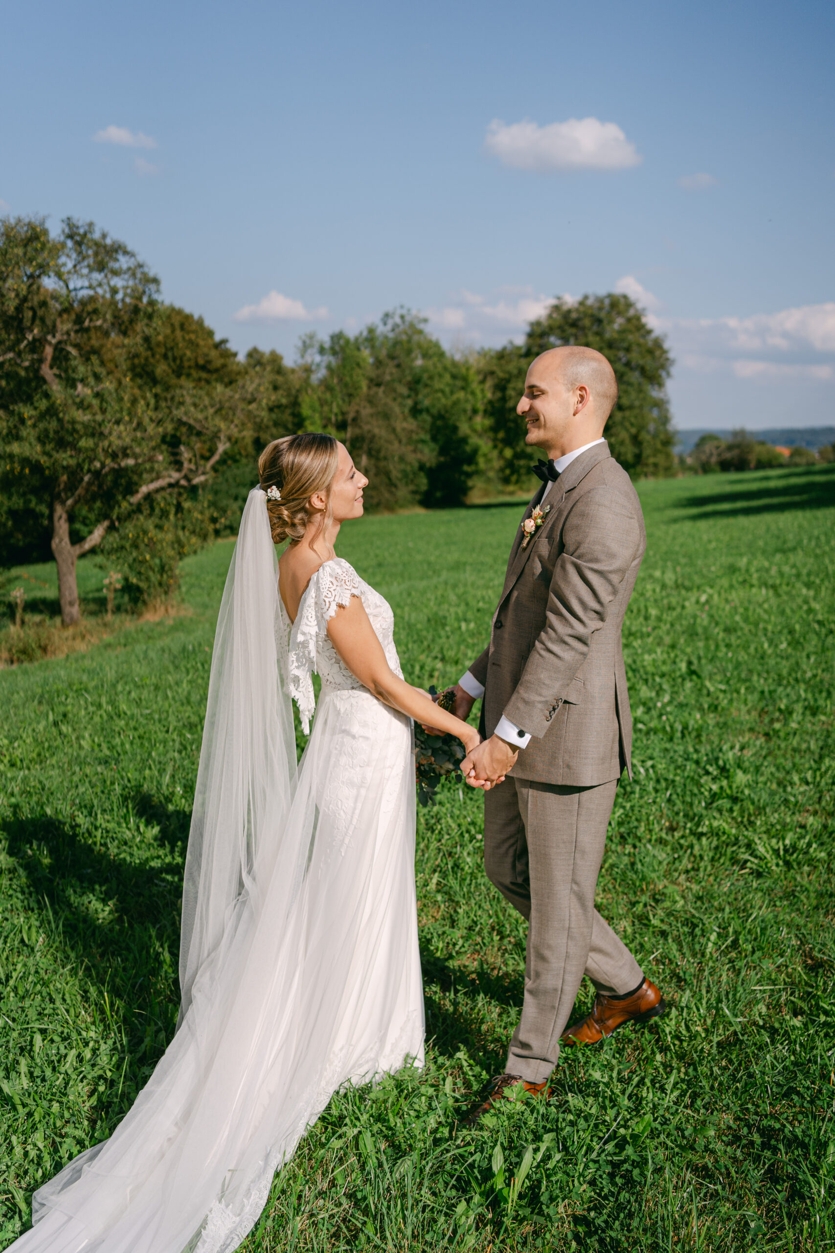 Hochzeitspaar im Sonnenlicht auf Wiese steht zueinander gewandt und halten Händchen. Sie schauen sich dabei liebevoll in die Augen.