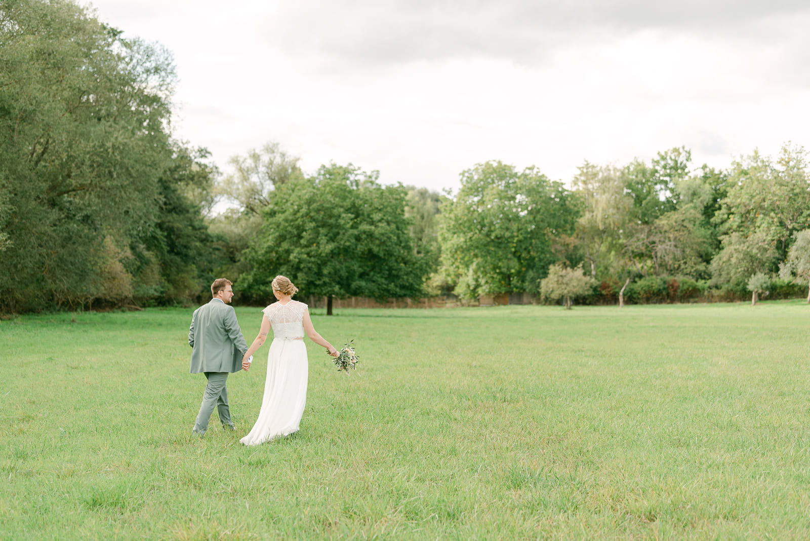 Hochzeitspaar auf Wiese läuft Händchen haltend von Kamera weg