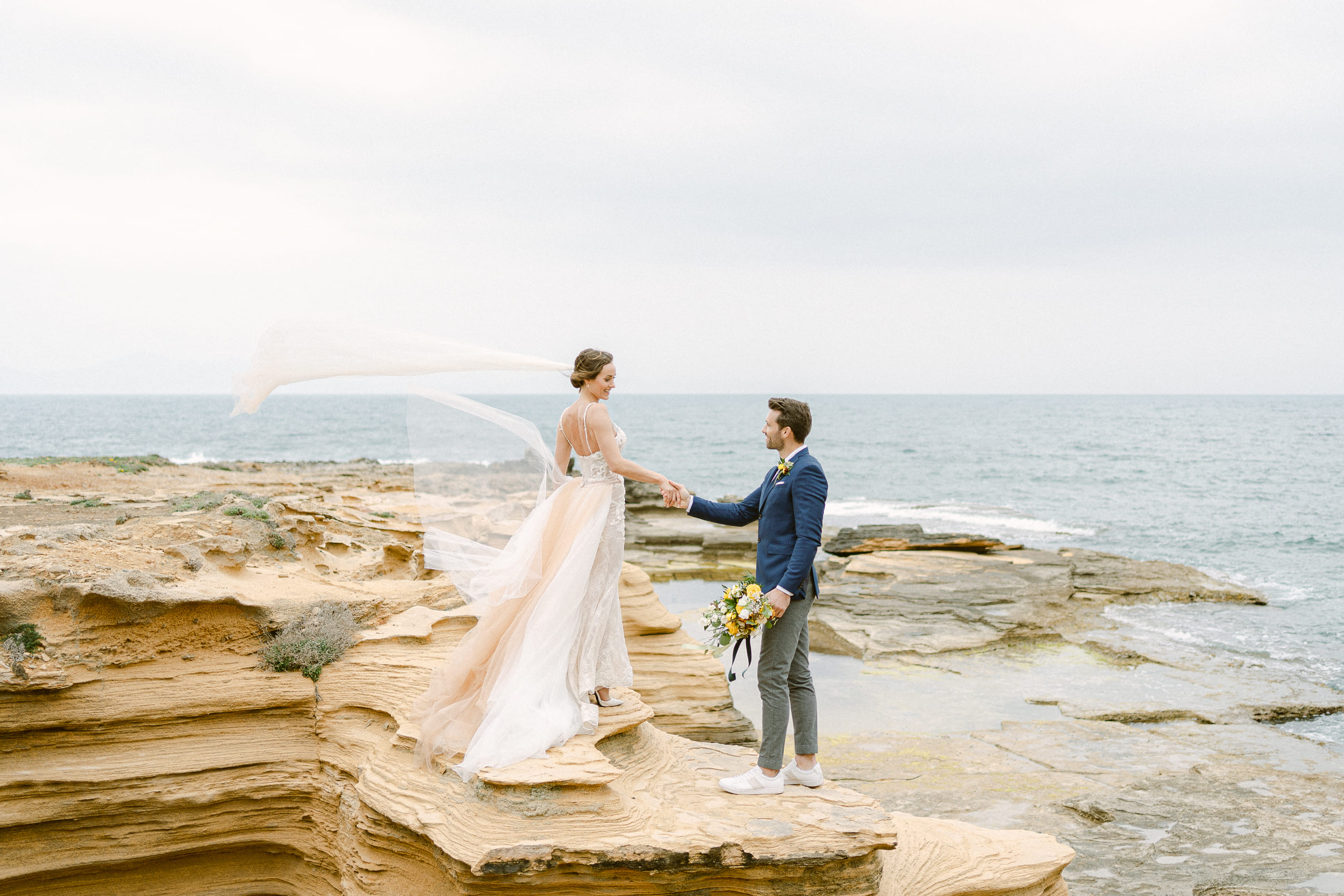 Hochzeitsfotograf Wiesbaden für eure wichtigsten Erinnerungen - Brautpaar steht auf Klippe am Meer, Wind weht Schleier in die Luft, das Paar hält Händchen
